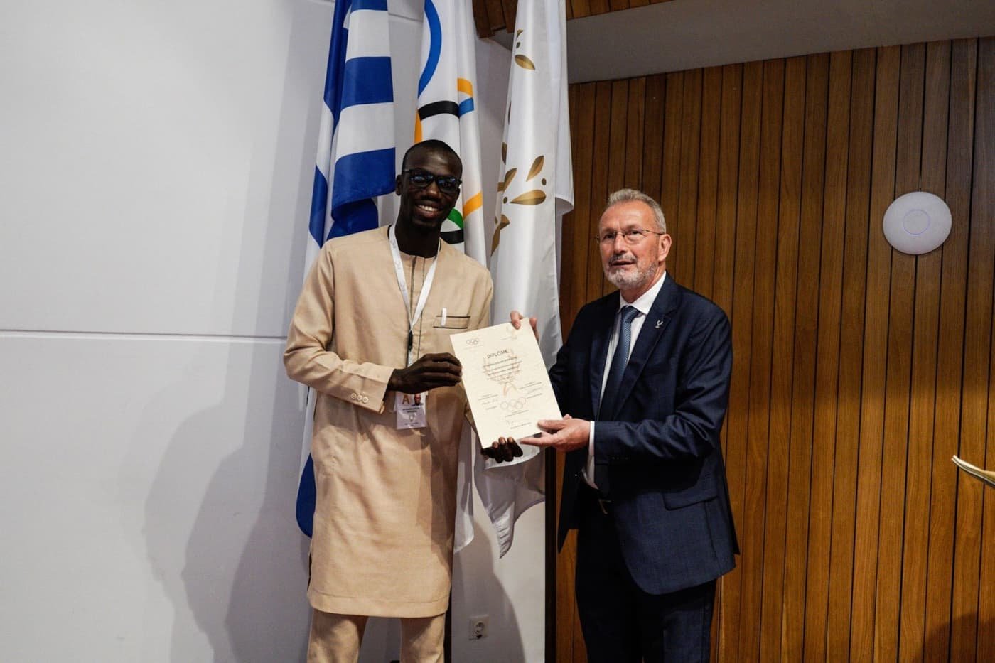 Remise de diplôme olympique devant drapeaux Grèce et olympique
